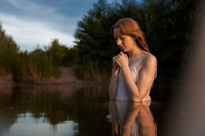Ve světle zapadajícího slunce se koupe rusovlasá dívka s dlouhými vlasy. Oblečena je do tenkých bílých šatů. Ruce má před sebou. Působí velmi křehce. Voda odráží obraz na klidné hladině.draz