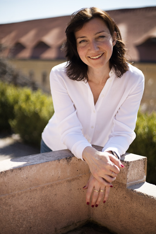 Elegantní dáma kolem 40 let, s mladistvým vzhledem se opírá o zídku. Bílá košile, polodlouhé tmavé vlasy. Opírá se uvolněně o zídku, ruce zkřížené přes zídku.