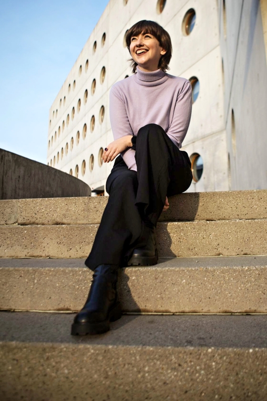 Mladá rozeskátá žena sedí na schoedech za Studijní a vědeckou knihovnou v Hradci Králové. Má lila rolám, černé kalhoty a černé bory s širokou podrážkou. Dívá se do dálky, ruce má zkřížené v klíně.