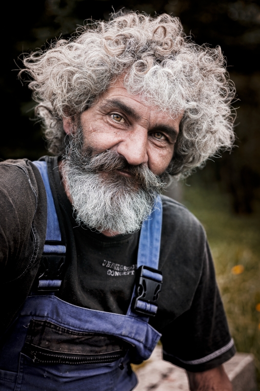 Muž v montérkách s bohatými kudrnatými šedivými vlasy a vousy se dívá do objektivu pronikavýma očima.