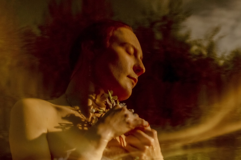 Na hladině se odráží dívka, drží v rukou květiny, tiskně je na prsa. Mokré vlasy má sčesané dozadu.