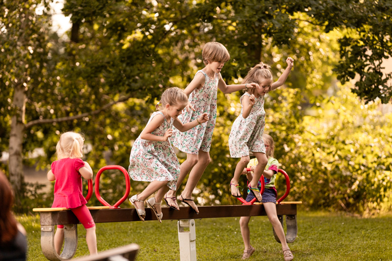 Pět holčiček řádí na kyvadlové houpačce. Dvě malé sedí na koncích a tři sestřičky ve stejných šatičkách skáče ze středové osy houpačky.