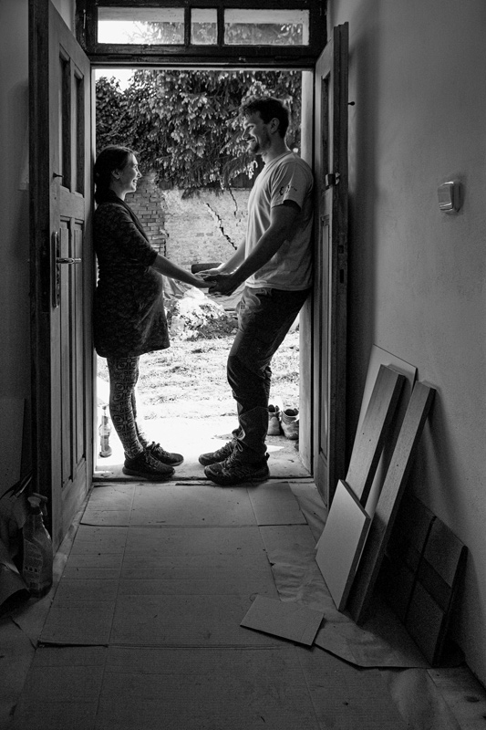 Mladý pár,  jsou v pracovním oblečení, ona je těhotná asi v 6. měsíci. Stojí ve dveřích svého rozestavěného domu, drží se za ruce, dívají se do očí a je cítit vzájemná láska. Černobílá fotografie.