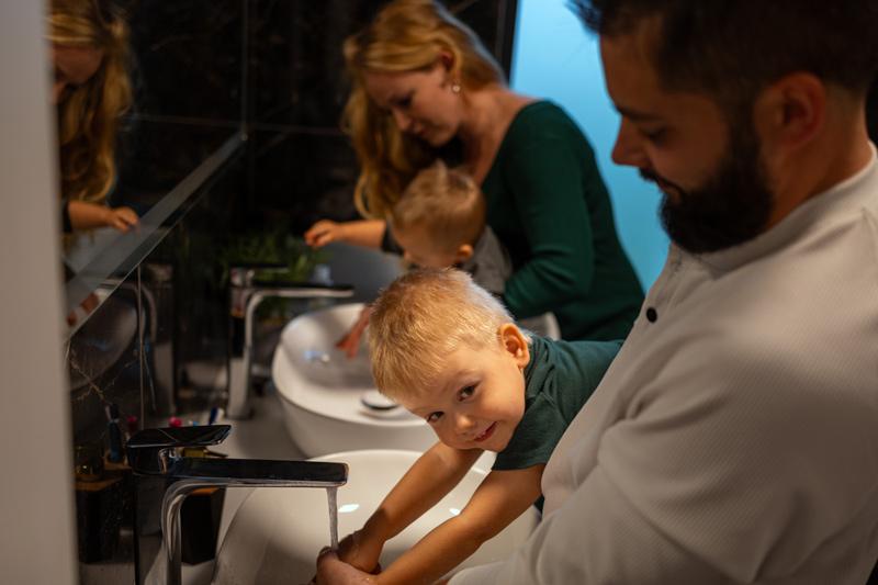 Rodiče pomáhají dvěma chlapččkům s mytím rukou. Tatínek s větším chlapcem jsou blíže k objektivu, maminka s menším jsou v pozadí. Děti jsou nakloněny nad umyvadly.