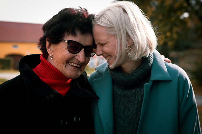 Starší dáma v tmavých brýlích a mladá žena blondýnka (Barbora Bočková) - babička a vnučka. Čelem se jedna druhé dotýkají a spiklenecky se smějí. Fotka má silný emoční náboj vzájemné lásky.
