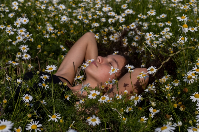 Mladá dívka leží v louce plné kvetoucích heřmánků. Ruce má pod hlavou, zavřené oči. Fotka sálá letní atmosféru..