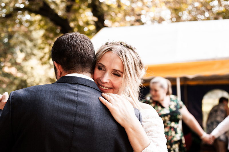 Ženich a nevsta kolem, asi čtyřicátníci si užívají svatební tanec. Nevěsta je opřená o rameno ženich a je jí vidět do obličeje. Je šťastná. Ženich je zády k objektivu. V pozadí tančí svatebčané v kruhu.