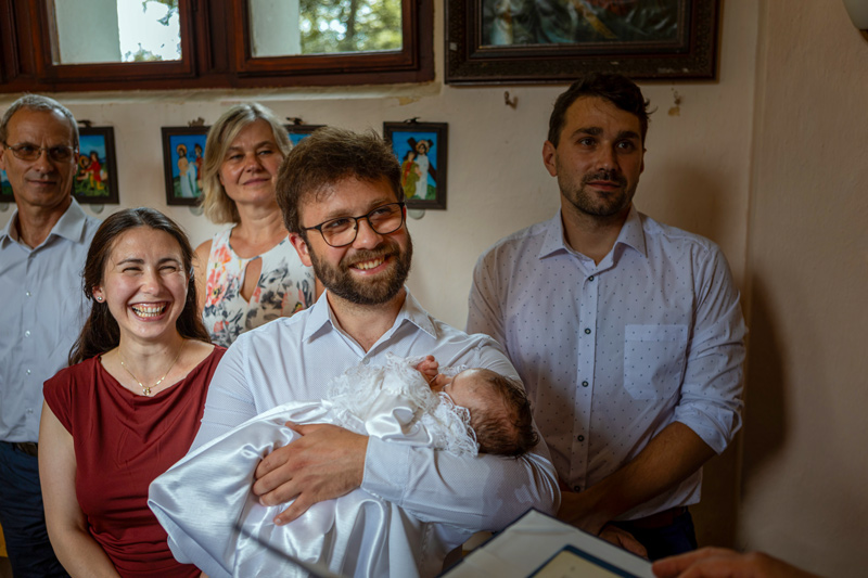 Křtiny v malé kapličce. Kmotr drží v náručí miminko, rodiče a prarodiče stojí v pozadí. Farář je zřejmě něčím právě rozesmál.